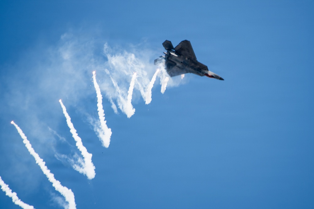 A stealth fighter jet, consistent with an F-22 Raptor, is captured in a steep banking maneuver against a clear blue sky. The aircraft is dispensing multiple flares in a sequential pattern, leaving bright burning trails and white smoke streaks across the frame. This appears to be an airshow demonstration showcasing the aircraft's countermeasure capabilities.