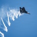 A stealth fighter jet, consistent with an F-22 Raptor, is captured in a steep banking maneuver against a clear blue sky. The aircraft is dispensing multiple flares in a sequential pattern, leaving bright burning trails and white smoke streaks across the frame. This appears to be an airshow demonstration showcasing the aircraft's countermeasure capabilities.