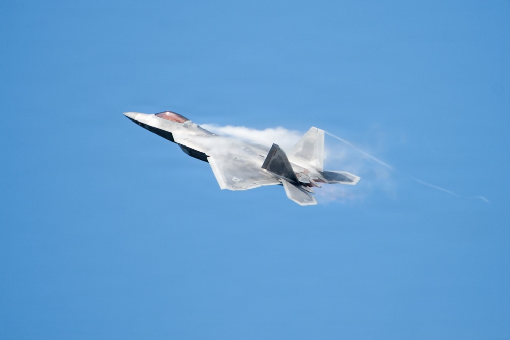 A fifth-generation stealth fighter jet, consistent with an F-22 Raptor, is captured in flight against a clear blue sky. The aircraft is banking steeply while generating visible vapor condensation trails along its fuselage and wing surfaces, indicative of high-speed, high-G maneuvering. The image appears to have been taken at an airshow or during a demonstration flight.