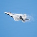 A fifth-generation stealth fighter jet, consistent with an F-22 Raptor, is captured in flight against a clear blue sky. The aircraft is banking steeply while generating visible vapor condensation trails along its fuselage and wing surfaces, indicative of high-speed, high-G maneuvering. The image appears to have been taken at an airshow or during a demonstration flight.