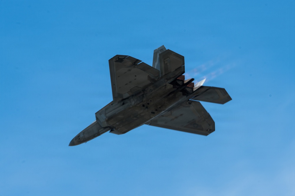 A stealth fighter jet, consistent with an F-22 Raptor, is photographed from below at a steep banking angle against a clear blue sky. The aircraft appears to be in afterburner, with visible exhaust flame and a short vapor trail emanating from the engines. The image captures the aircraft's distinctive diamond-shaped planform and angular stealth geometry.