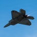 A stealth fighter jet, consistent with an F-22 Raptor, is photographed from below at a steep banking angle against a clear blue sky. The aircraft appears to be in afterburner, with visible exhaust flame and a short vapor trail emanating from the engines. The image captures the aircraft's distinctive diamond-shaped planform and angular stealth geometry.