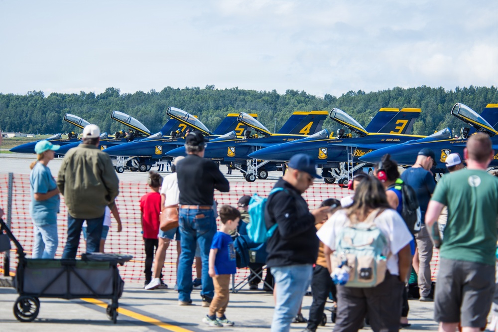 An air show event featuring the U.S. Navy Blue Angels flight demonstration squadron, with multiple F/A-18 Hornet jets numbered 3, 4, 5, and others parked on the tarmac. A large crowd of spectators, including families with children, are gathered behind orange safety fencing to view the aircraft up close. The event appears to be a public open house or static display at a military airfield.