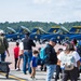 An air show event featuring the U.S. Navy Blue Angels flight demonstration squadron, with multiple F/A-18 Hornet jets numbered 3, 4, 5, and others parked on the tarmac. A large crowd of spectators, including families with children, are gathered behind orange safety fencing to view the aircraft up close. The event appears to be a public open house or static display at a military airfield.