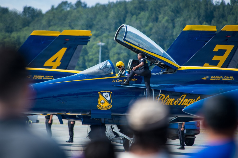 The image shows Blue Angels Boeing F/A-18 Super Hornet jets numbered 3 and 4 on a tarmac during what appears to be an airshow. A pilot wearing a yellow helmet is seated in the cockpit of jet number 4, while a crew member in a dark uniform assists from outside. Spectators are visible in the foreground watching the event.