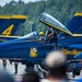 The image shows Blue Angels Boeing F/A-18 Super Hornet jets numbered 3 and 4 on a tarmac during what appears to be an airshow. A pilot wearing a yellow helmet is seated in the cockpit of jet number 4, while a crew member in a dark uniform assists from outside. Spectators are visible in the foreground watching the event.
