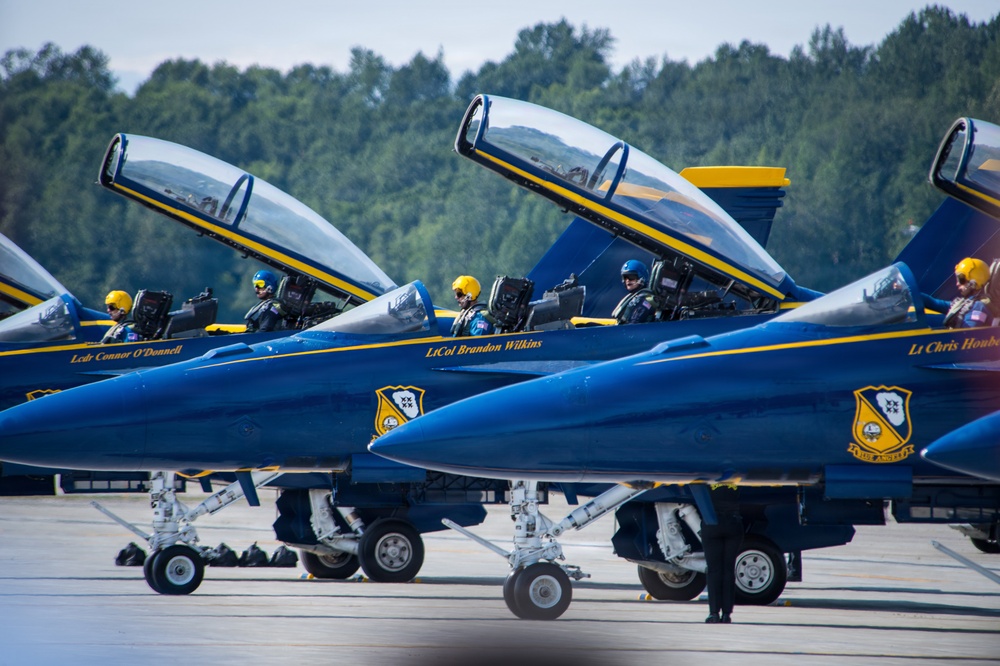 A line of U.S. Navy Blue Angels F/A-18 Super Hornet aircraft are staged on a tarmac with canopies open and pilots seated in cockpits, appearing to be preparing for a performance or taxi sequence. The jets display their iconic blue and gold color scheme along with individual pilot name placards on the fuselage. At least three aircraft are clearly visible with readable pilot names including LtCol Brandon Wilkins and LCdr Connor O'Donnell.