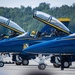 A line of U.S. Navy Blue Angels F/A-18 Super Hornet aircraft are staged on a tarmac with canopies open and pilots seated in cockpits, appearing to be preparing for a performance or taxi sequence. The jets display their iconic blue and gold color scheme along with individual pilot name placards on the fuselage. At least three aircraft are clearly visible with readable pilot names including LtCol Brandon Wilkins and LCdr Connor O'Donnell.