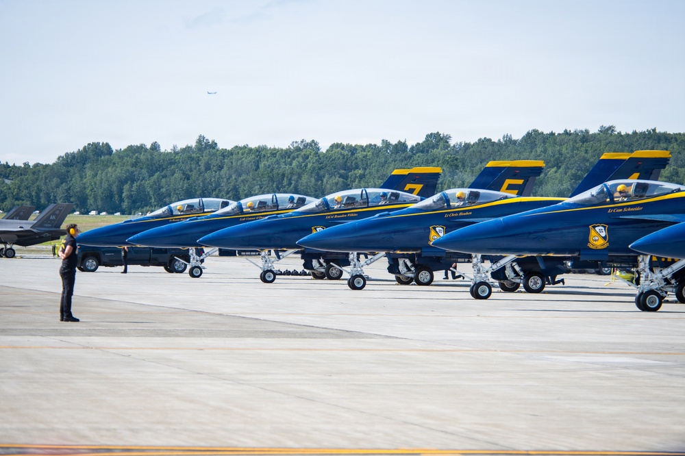 A line of U.S. Navy Blue Angels aircraft are parked on a tarmac at an airshow or military airfield. The jets are painted in their iconic blue and gold livery and are numbered, with at least numbers 4, 5, 6, and 7 visible on the tail fins. A ground crew member wearing ear protection stands on the tarmac observing the aircraft.