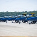 A line of U.S. Navy Blue Angels aircraft are parked on a tarmac at an airshow or military airfield. The jets are painted in their iconic blue and gold livery and are numbered, with at least numbers 4, 5, 6, and 7 visible on the tail fins. A ground crew member wearing ear protection stands on the tarmac observing the aircraft.