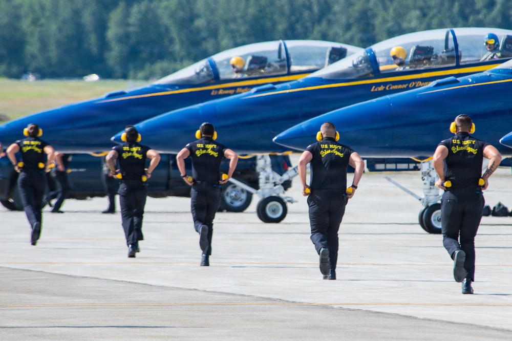 The image shows U.S. Navy Blue Angels ground crew members running across a tarmac toward a line of Blue Angels F/A-18 jets preparing for takeoff. The crew members are dressed in matching black uniforms with yellow 'U.S. Navy Blue Angels' lettering on the back and are wearing yellow ear protection. At least two jets are visible with pilots seated in the cockpits, and pilot name placards are visible on the fuselages.