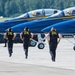 The image shows U.S. Navy Blue Angels ground crew members running across a tarmac toward a line of Blue Angels F/A-18 jets preparing for takeoff. The crew members are dressed in matching black uniforms with yellow 'U.S. Navy Blue Angels' lettering on the back and are wearing yellow ear protection. At least two jets are visible with pilots seated in the cockpits, and pilot name placards are visible on the fuselages.