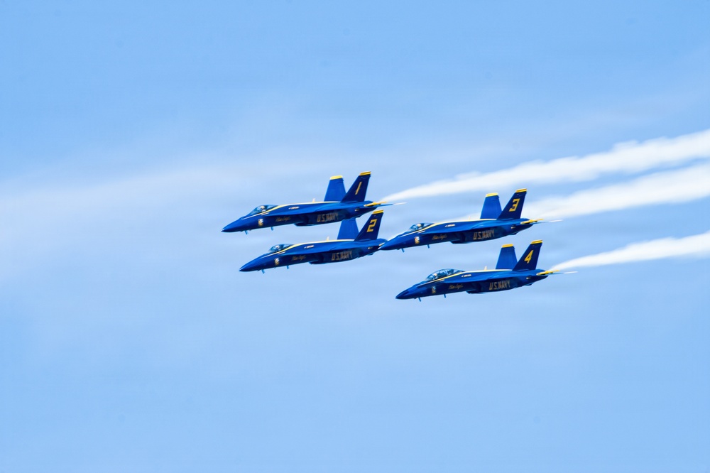 Four U.S. Navy Blue Angels aircraft are flying in a tight diamond formation against a clear blue sky. The jets, numbered 1 through 4, are painted in their signature blue and gold livery with 'U.S. NAVY' markings. White contrails stream behind the aircraft, emphasizing their speed and precision formation flying.