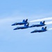 Four U.S. Navy Blue Angels aircraft are flying in a tight diamond formation against a clear blue sky. The jets, numbered 1 through 4, are painted in their signature blue and gold livery with 'U.S. NAVY' markings. White contrails stream behind the aircraft, emphasizing their speed and precision formation flying.