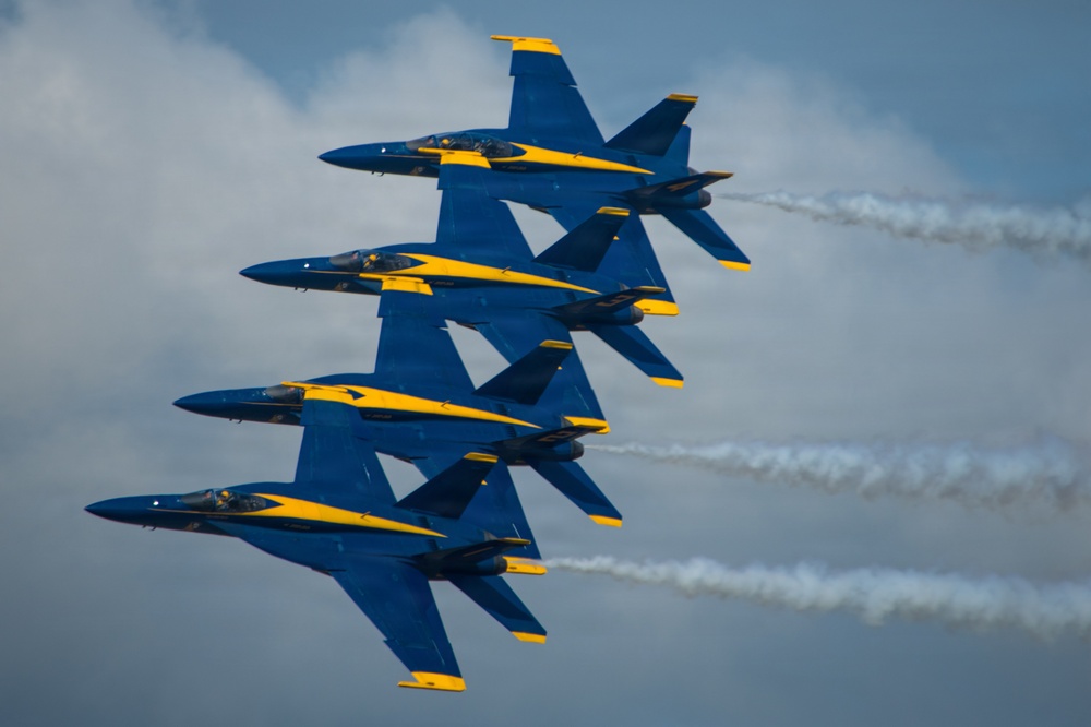 Four military jet aircraft painted in blue and yellow livery fly in a tight diagonal formation, each trailing white smoke streams. The aircraft are consistent with the U.S. Navy Blue Angels flight demonstration squadron, flying in their signature close-formation diamond or echelon configuration. The image is captured from a close angle against a partly cloudy sky, emphasizing the precision and proximity of the formation.