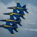 Four military jet aircraft painted in blue and yellow livery fly in a tight diagonal formation, each trailing white smoke streams. The aircraft are consistent with the U.S. Navy Blue Angels flight demonstration squadron, flying in their signature close-formation diamond or echelon configuration. The image is captured from a close angle against a partly cloudy sky, emphasizing the precision and proximity of the formation.