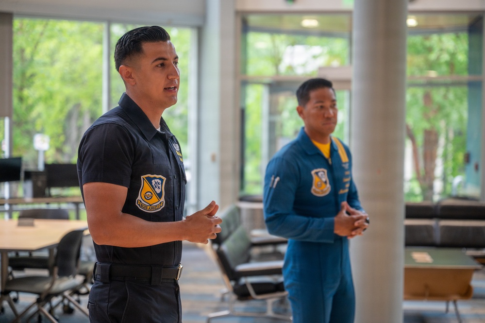Two individuals wearing Blue Angels flight team uniforms are standing in what appears to be a modern indoor facility, likely during a public appearance or presentation. The person in the foreground is speaking while gesturing with their hands, while the second person stands attentively in the background. Both wear distinctive Blue Angels patches on their uniforms.