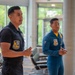 Two individuals wearing Blue Angels flight team uniforms are standing in what appears to be a modern indoor facility, likely during a public appearance or presentation. The person in the foreground is speaking while gesturing with their hands, while the second person stands attentively in the background. Both wear distinctive Blue Angels patches on their uniforms.