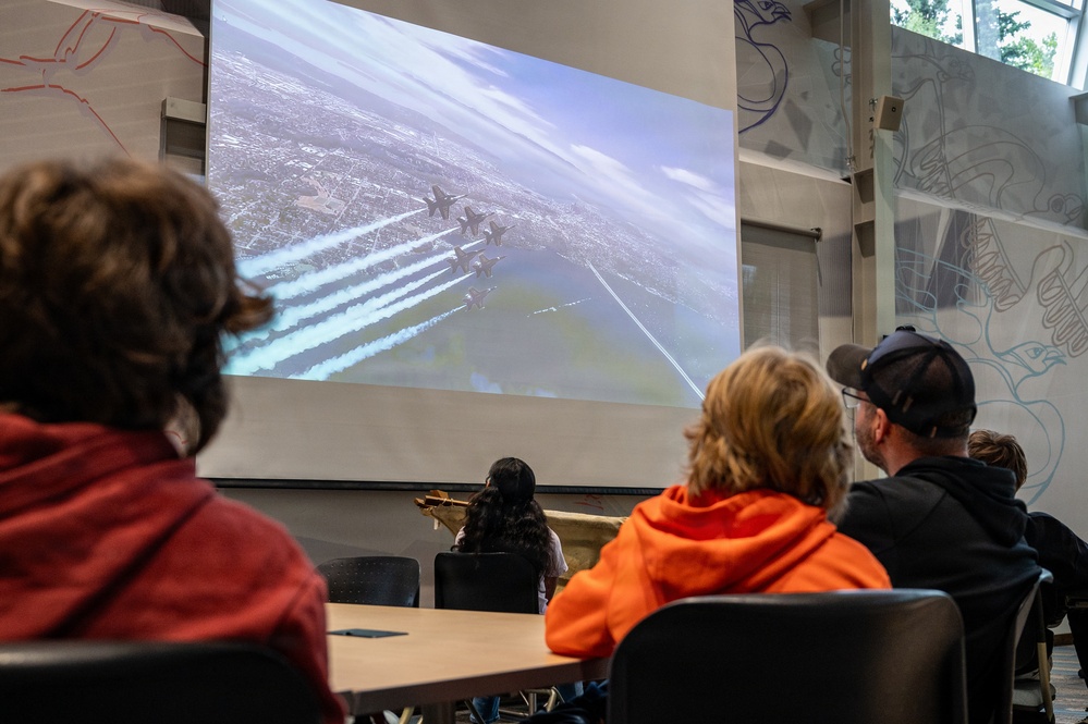 A group of people are seated at tables in what appears to be a classroom or presentation room, watching a large projected screen. The screen displays an aerial formation of military jets, likely the Blue Angels, flying in tight formation over a cityscape with visible smoke trails. The setting suggests an educational or informational presentation about aviation or military aerobatics.