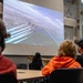 A group of people are seated at tables in what appears to be a classroom or presentation room, watching a large projected screen. The screen displays an aerial formation of military jets, likely the Blue Angels, flying in tight formation over a cityscape with visible smoke trails. The setting suggests an educational or informational presentation about aviation or military aerobatics.