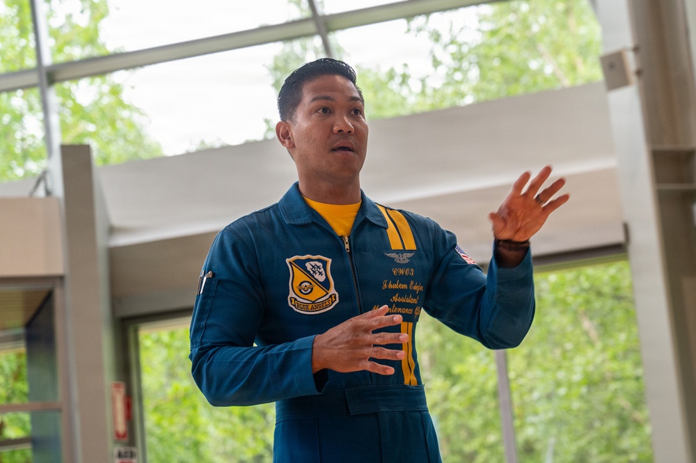 A U.S. Navy Blue Angels team member in a blue and gold flight suit is speaking or presenting, gesturing with both hands. The patch on his chest reads 'Blue Angels' and his name tag indicates the rank of CWO3 with the title 'Assistant Maintenance Officer.' He appears to be in an indoor facility with large windows and green trees visible in the background.