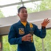 A U.S. Navy Blue Angels team member in a blue and gold flight suit is speaking or presenting, gesturing with both hands. The patch on his chest reads 'Blue Angels' and his name tag indicates the rank of CWO3 with the title 'Assistant Maintenance Officer.' He appears to be in an indoor facility with large windows and green trees visible in the background.