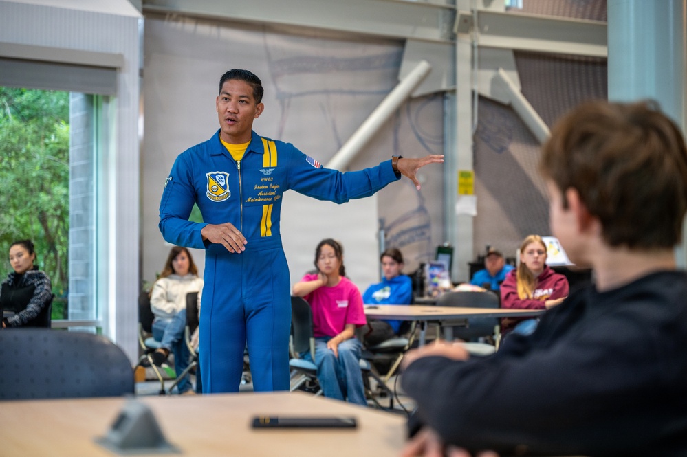 A person wearing a blue U.S. Navy Blue Angels flight suit is presenting or speaking to a group of students in what appears to be a classroom or educational facility. The speaker is gesturing with one hand while addressing the seated audience. The setting suggests an educational outreach or community engagement event.