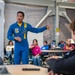 A person wearing a blue U.S. Navy Blue Angels flight suit is presenting or speaking to a group of students in what appears to be a classroom or educational facility. The speaker is gesturing with one hand while addressing the seated audience. The setting suggests an educational outreach or community engagement event.
