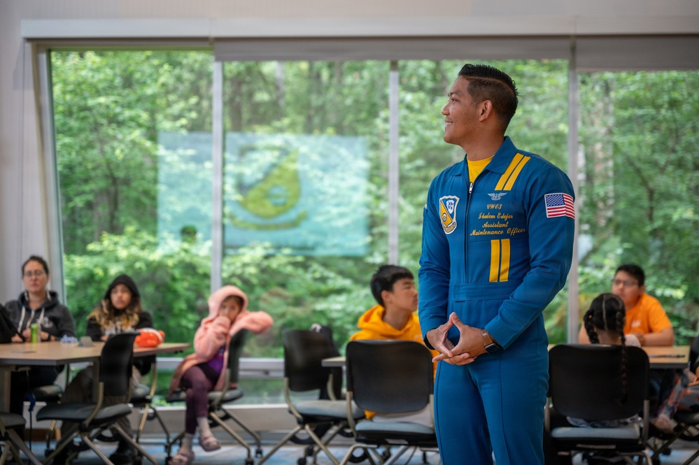 A U.S. Navy Blue Angels crew member in a distinctive blue flight suit is presenting or speaking to a group of young people in what appears to be a classroom or educational facility. The speaker's name tag reads 'Shalon Edger, Assistant Maintenance Officer' and he is wearing the iconic Blue Angels uniform with yellow accents and a U.S. flag patch. The audience, consisting of children and young adults, is seated at tables in a room with large windows overlooking a wooded area.
