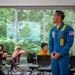A U.S. Navy Blue Angels crew member in a distinctive blue flight suit is presenting or speaking to a group of young people in what appears to be a classroom or educational facility. The speaker's name tag reads 'Shalon Edger, Assistant Maintenance Officer' and he is wearing the iconic Blue Angels uniform with yellow accents and a U.S. flag patch. The audience, consisting of children and young adults, is seated at tables in a room with large windows overlooking a wooded area.