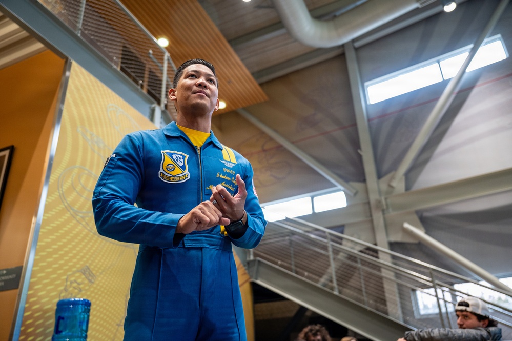 A person wearing a blue and gold flight suit associated with the U.S. Navy Blue Angels demonstration squadron is standing in what appears to be a modern indoor facility. The individual appears to be speaking or presenting to an audience, with hands clasped together in a conversational gesture. The setting suggests a public outreach or community engagement event.