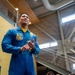 A person wearing a blue and gold flight suit associated with the U.S. Navy Blue Angels demonstration squadron is standing in what appears to be a modern indoor facility. The individual appears to be speaking or presenting to an audience, with hands clasped together in a conversational gesture. The setting suggests a public outreach or community engagement event.