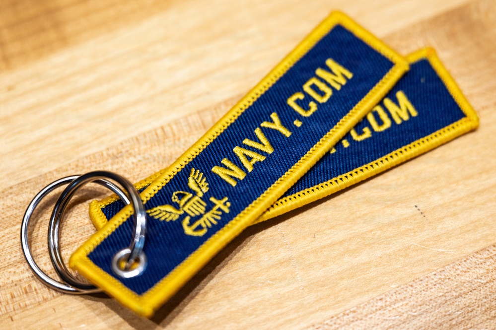 The image shows a embroidered fabric keychain tag with 'NAVY.COM' text in gold lettering on a navy blue background with gold border. The keychain features a U.S. Navy eagle insignia embroidered below the text and is attached to a silver split ring. Two overlapping tags are visible, resting on a light wood surface.