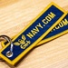 The image shows a embroidered fabric keychain tag with 'NAVY.COM' text in gold lettering on a navy blue background with gold border. The keychain features a U.S. Navy eagle insignia embroidered below the text and is attached to a silver split ring. Two overlapping tags are visible, resting on a light wood surface.