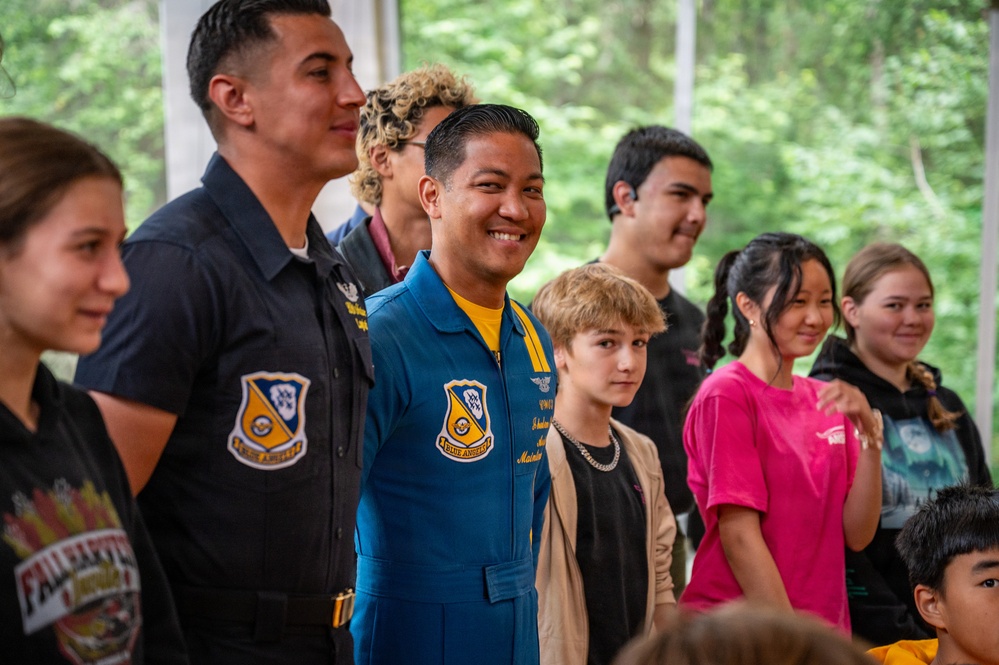 A group of people, including at least two individuals wearing Blue Angels flight suits and uniforms, pose together in what appears to be an outdoor or semi-outdoor setting with trees visible in the background. The central figure is smiling broadly and wearing a blue and yellow Blue Angels flight suit with visible insignia. The gathering appears to be a meet-and-greet or community outreach event involving youth and Blue Angels personnel.