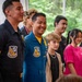 A group of people, including at least two individuals wearing Blue Angels flight suits and uniforms, pose together in what appears to be an outdoor or semi-outdoor setting with trees visible in the background. The central figure is smiling broadly and wearing a blue and yellow Blue Angels flight suit with visible insignia. The gathering appears to be a meet-and-greet or community outreach event involving youth and Blue Angels personnel.