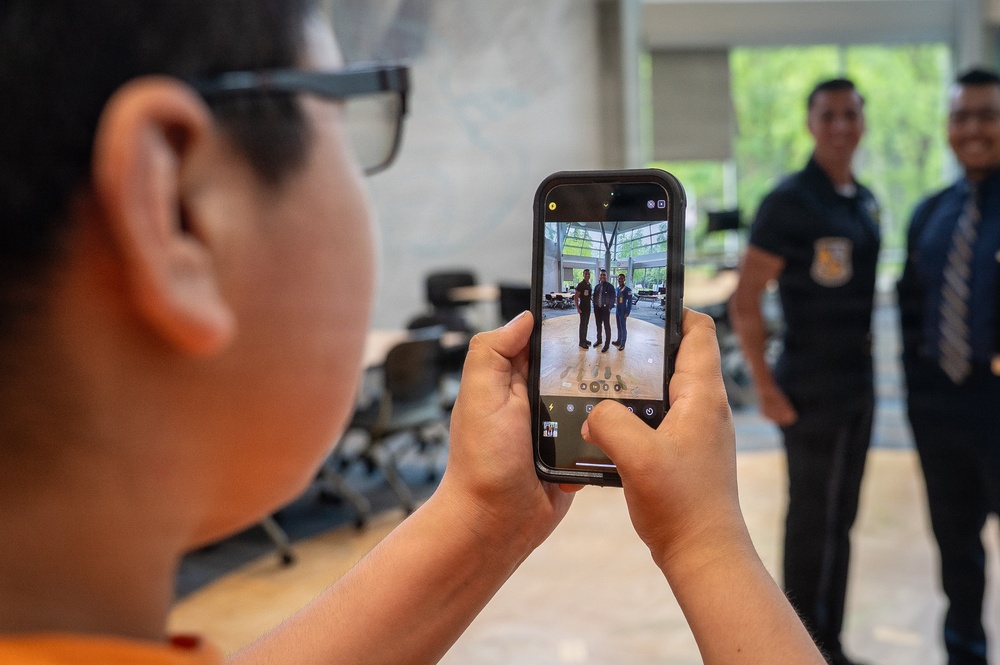 A young child wearing glasses and an orange shirt is photographing two uniformed men using a smartphone with a black case. The two men in the background are smiling and posing for the photo, wearing dark uniforms with insignia patches. The smartphone screen clearly shows the live camera preview of the two subjects being photographed.