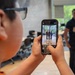 A young child wearing glasses and an orange shirt is photographing two uniformed men using a smartphone with a black case. The two men in the background are smiling and posing for the photo, wearing dark uniforms with insignia patches. The smartphone screen clearly shows the live camera preview of the two subjects being photographed.
