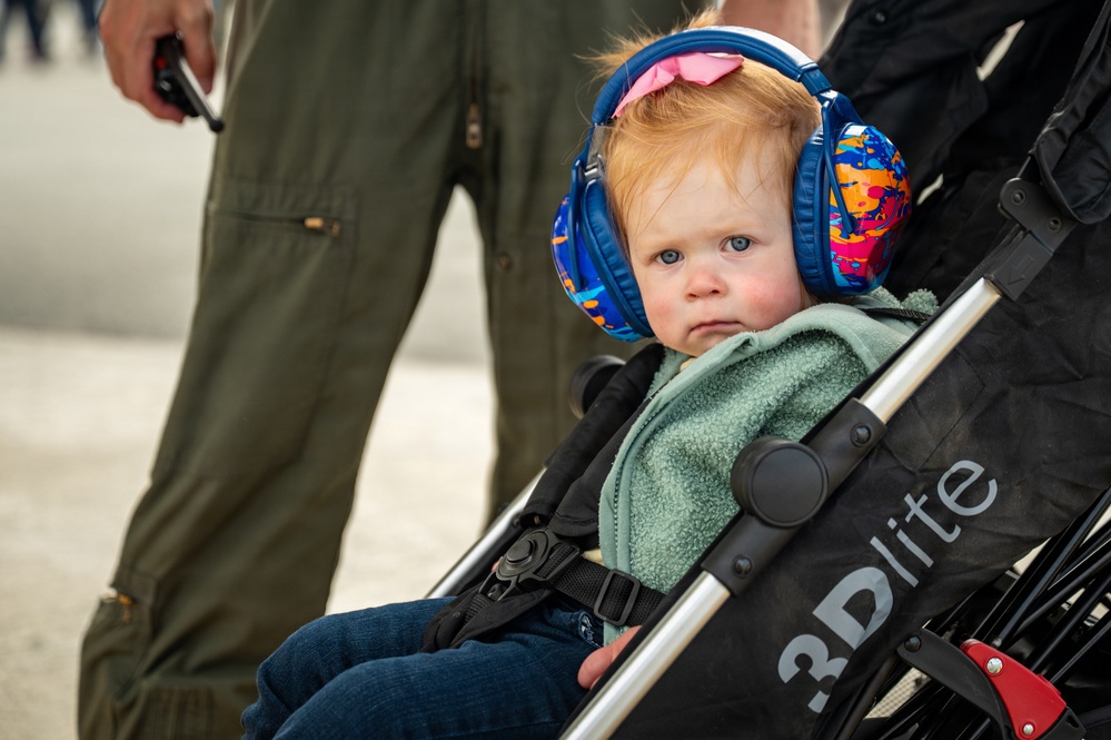 A young toddler with red hair and blue eyes sits in a '3Dlite' brand stroller, wearing large blue noise-canceling/hearing protection earmuffs with a colorful pattern and a pink bow on top. The child is dressed in a mint green fleece jacket and jeans, appearing to be at an outdoor event, likely an airshow given the adult in the background wearing an olive green flight suit. The child's expression is calm but slightly serious.