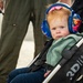 A young toddler with red hair and blue eyes sits in a '3Dlite' brand stroller, wearing large blue noise-canceling/hearing protection earmuffs with a colorful pattern and a pink bow on top. The child is dressed in a mint green fleece jacket and jeans, appearing to be at an outdoor event, likely an airshow given the adult in the background wearing an olive green flight suit. The child's expression is calm but slightly serious.