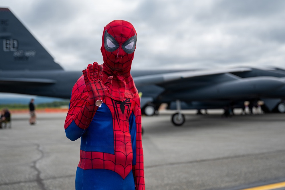 A person dressed in a full Spider-Man costume poses on a military airfield tarmac, raising one hand in a web-shooting gesture. Behind them is a large military aircraft, identifiable by partial tail markings reading 'ED', consistent with Edwards Air Force Base. The setting appears to be an air show or public event, with other people visible in the background.