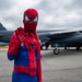 A person dressed in a full Spider-Man costume poses on a military airfield tarmac, raising one hand in a web-shooting gesture. Behind them is a large military aircraft, identifiable by partial tail markings reading 'ED', consistent with Edwards Air Force Base. The setting appears to be an air show or public event, with other people visible in the background.