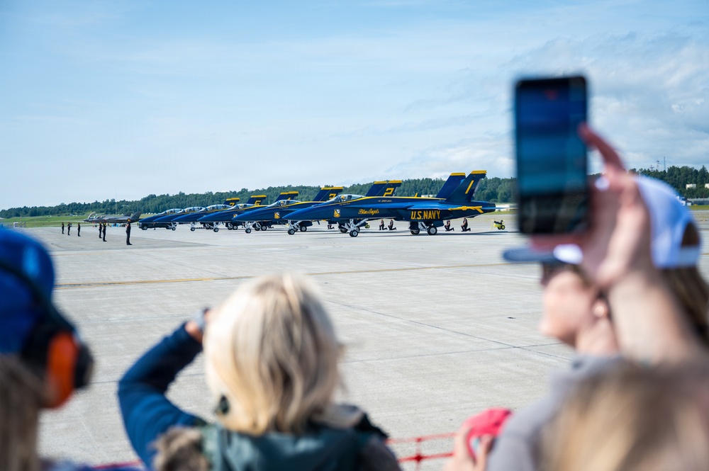 A crowd of spectators watches and photographs a lineup of U.S. Navy Blue Angels aircraft on an airfield tarmac. The jets, painted in their distinctive blue and gold livery, are numbered and arranged in a row, with ground crew and personnel visible nearby. The scene appears to be an air show or public demonstration event on a clear, sunny day.