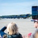 A crowd of spectators watches and photographs a lineup of U.S. Navy Blue Angels aircraft on an airfield tarmac. The jets, painted in their distinctive blue and gold livery, are numbered and arranged in a row, with ground crew and personnel visible nearby. The scene appears to be an air show or public demonstration event on a clear, sunny day.