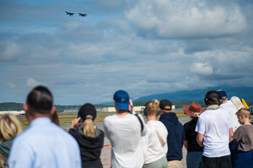 A crowd of spectators stands along an airfield perimeter watching two military jet aircraft flying in close formation overhead. The scene appears to be an airshow event, with mountains and runway infrastructure visible in the background. The foreground crowd is intentionally blurred using shallow depth of field, keeping focus on the aircraft in the sky.