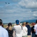 A crowd of spectators stands along an airfield perimeter watching two military jet aircraft flying in close formation overhead. The scene appears to be an airshow event, with mountains and runway infrastructure visible in the background. The foreground crowd is intentionally blurred using shallow depth of field, keeping focus on the aircraft in the sky.