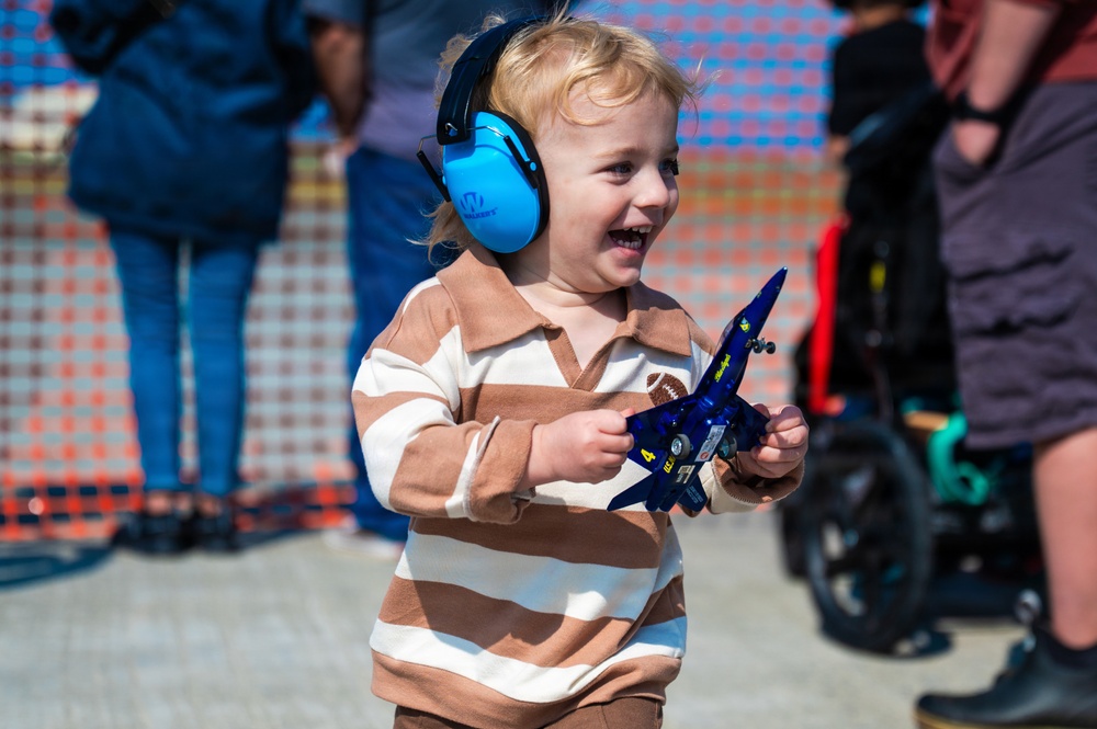 A young toddler with blonde hair is smiling and holding a blue toy jet aircraft, likely a Blue Angels F/A-18 model. The child is wearing bright blue noise-canceling ear protection headphones, suggesting they are at a loud outdoor event such as an airshow. The child appears happy and engaged, dressed in a brown and white striped long-sleeve shirt.