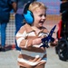A young toddler with blonde hair is smiling and holding a blue toy jet aircraft, likely a Blue Angels F/A-18 model. The child is wearing bright blue noise-canceling ear protection headphones, suggesting they are at a loud outdoor event such as an airshow. The child appears happy and engaged, dressed in a brown and white striped long-sleeve shirt.