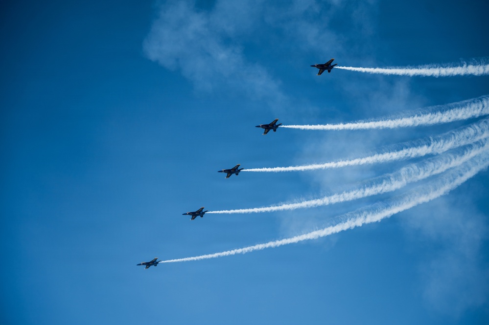 Five military jet aircraft fly in a diagonal echelon formation across a clear blue sky, each producing a distinct white smoke trail. The aircraft appear to be performing an aerobatic demonstration, consistent with a precision flight team display. The smoke trails fan out in parallel lines, creating a visually striking pattern against the bright sky.