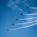 Five military jet aircraft fly in a diagonal echelon formation across a clear blue sky, each producing a distinct white smoke trail. The aircraft appear to be performing an aerobatic demonstration, consistent with a precision flight team display. The smoke trails fan out in parallel lines, creating a visually striking pattern against the bright sky.